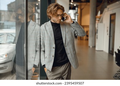 Focused male manager in eyeglasses is talking phone standing in modern coworking space - Shutterstock ID 2395459507