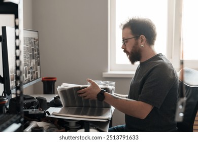 A focused male photographer examines printed photographs while sitting at a cluttered desk in a home office setup. - Powered by Shutterstock