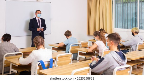 Focused male teacher in protective face mask giving lesson to teenage students in college. New life reality during coronavirus pandemic