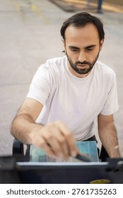 A focused man with a beard and nose ring is creating art using watercolors and a pen outside