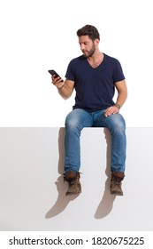 Focused Man In Boots, Jeans And Unbuttoned Lumberjack Shirt Is Sitting On A Top, Holding Telephone And Reading. Full Length Studio Shot Isolated On White.