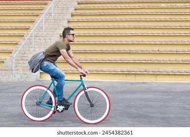 Focused man in casual attire rides a fixed-gear bike in an urban setting, with a messenger bag over his shoulder, portraying active, eco-friendly transportation - Powered by Shutterstock