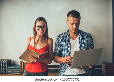 Focused man in checkered shirt typing on laptop while female friend in glasses dressed in red tank top writing in notepad smiling and looking at camera in library - Powered by Shutterstock