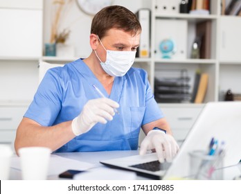 Focused Man Doctor Wearing Medical Facial Mask And Gloves Working In Medical Office Using Laptop Computer