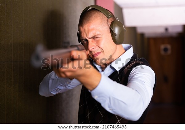 Focused man in ear protectors aiming\
single barrelled shotgun at target in shooting\
range.