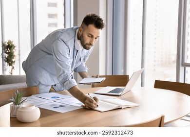 Focused man multitasking, working on analytical task, prepare presentation or conducting research standing at table, holds copybook while writes, laptop, printed documents spread across workplace desk