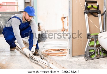 Focused man running cable and conduit through concrete trench at indoor construction site