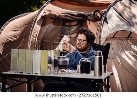 Focused man sets up a folding windshield for a camp stove outside a tent, surrounded by outdoor cooking gear on a table.
