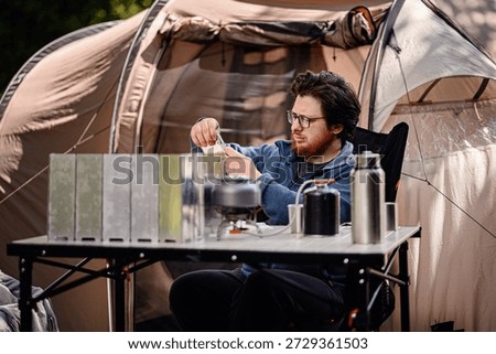 Focused man sets up a folding windshield for a camp stove outside a tent, surrounded by outdoor cooking gear on a table.