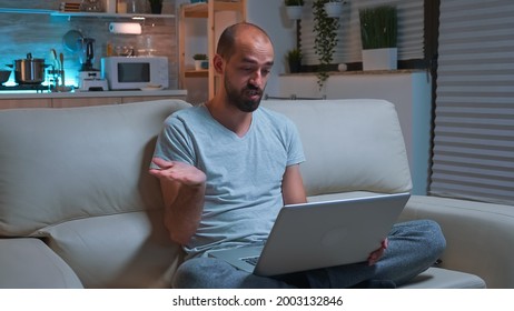 Focused Man Sitting On Sofa In Pajamas While Talking With Collegue During Videocall Conference Using Laptop Computer. Caucasian Male Working At Communication Project Late At Night In Kitchen