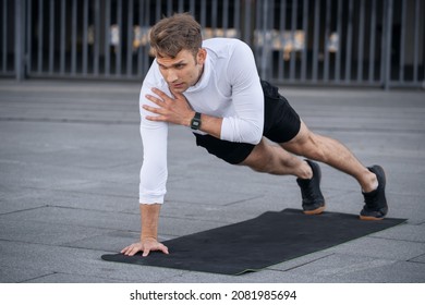 Focused man in sportswear doing shoulder tap in push or press ups exercise, standing in plank pose. Side view of confident and serious sportsman with smart watch training outdoor, work out on yoga mat