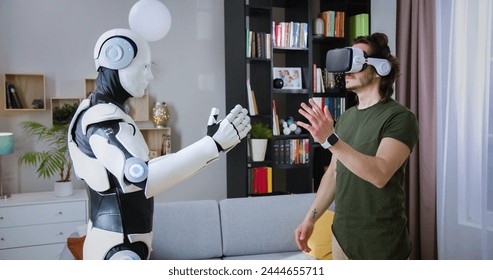 Focused man in virtual reality headset examining robot's movements in bright living room. Humanoid cyborg giving high five to male in casual wear and goggles.
