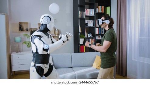 Focused man in virtual reality headset examining robot's movements in bright living room. Humanoid cyborg giving high five to male in casual wear and goggles.