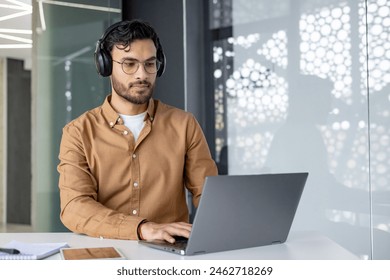 A focused man wearing headphones and glasses, working on a laptop in a modern office setting. He is concentrated on his task with a notepad and tablet on the table. - Powered by Shutterstock