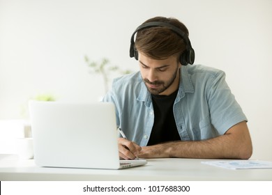 Focused Man Wearing Headphones Writing Notes Studying With Laptop, Serious Student Or Office Worker Listening To Audio Business Course At Work, E-learning And Online Professional Education Concept