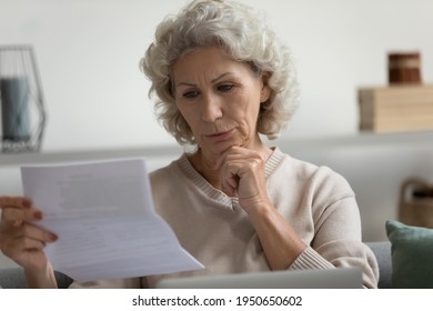 Focused Mature 60s Retired Lady Receiving Letter And Reading Document At Home. Serious Middle Aged Woman Checking Bank Loan Notification, Tax Form, Making Online Payment Of Bill Or Invoice