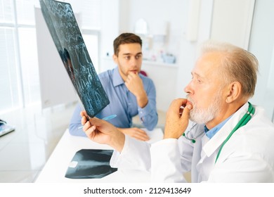 Focused Mature Adult Male Physician Consult Frustrated Young Man Patient Giving Bad News Explaining Results Of MRI Image. Unhappy Scared Young Man Listening To Bad News Sitting In Doctor Office.