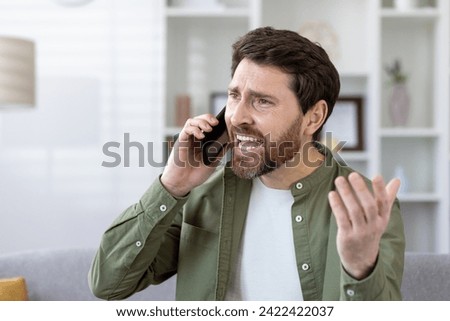 Focused mature bearded man in a casual shirt having a conversation on smartphone at his well-lit living room.