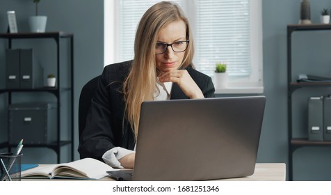 Focused mature business lady in formal suit and specs working on laptop sitting at office table