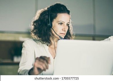 Focused mature call center operator looking at laptop. Serious curly woman working in office. Call center concept