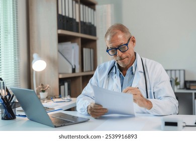 Focused mature doctor reviewing patient files, working on laptop in medical office, demonstrating professionalism and healthcare expertise - Powered by Shutterstock