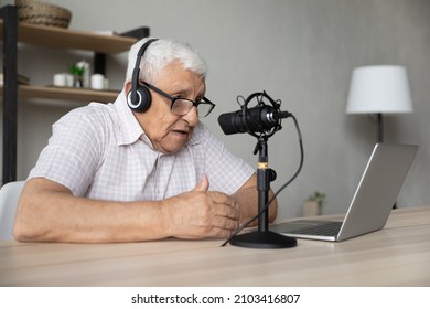 Focused mature elderly retired man in eyeglasses wearing wireless headphones looking at computer screen, talking in professional stand microphone, voice acting or recording vlog alone at home.