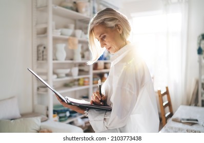 Focused mature european female entrepreneur writing something in clipboard. Small business and entrepreneurship. Modern successful woman. Home art studio with pottery on shelves at sunny day