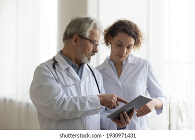 Focused mature male doctor and female nurse look at tablet screen discuss anamnesis together. Concentrated diverse medical professionals use pad device, engaged in team thinking in hospital.