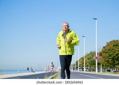 Focused Mature Man In Green Sports Jacket And Tights Jogging Along River Bank Outside. Senior Jogger Training For Marathon. Front View. Activity And Age Concept