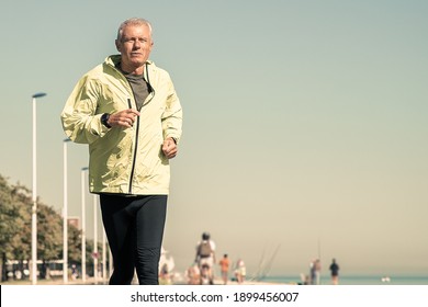 Focused Mature Man In Green Sports Jacket And Tights Jogging Along River Bank Outside. Senior Jogger Training For Marathon. Front View. Activity And Age Concept