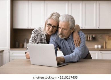 Focused mature spouses using laptop in the kitchen, learn commercial offer, reading received e-mail from bank, making purchases using delivery services. Older generation and modern tech usage at home - Powered by Shutterstock