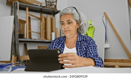 A focused mature woman carpenter using tablet in a well-lit workshop with woodworking tools. - Powered by Shutterstock