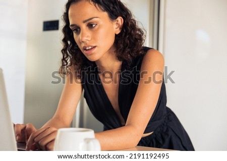 Focused mid adult female freelancer in black attire using laptop by coffee cup on kitchen island at home