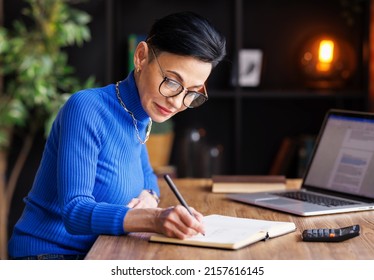 Focused middle aged female entrepreneur working at laptop and take notes  while working on project in domestic workplace - Powered by Shutterstock