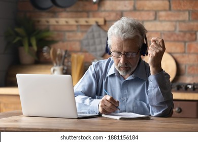 Focused Middle Aged Mature Man In Glasses Wearing Headphones, Involved In Studying On Online Courses Using Computer Applications. Concentrated Older Grandfather Watching Online Webinar, Writing Notes.