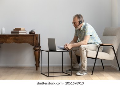 Focused Middle Aged Old Man In Glasses Wearing Headset With Microphone, Listening Educational Lecture, Studying Distantly On Online Courses On Computer, Improving Knowledge Writing Notes In Copybook.