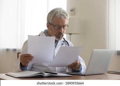 Focused middle aged senior male general practitioner in eyewear looking through paper documents, reading medical insurance contracts, reviewing reports or illness treatment history at clinic. - Powered by Shutterstock