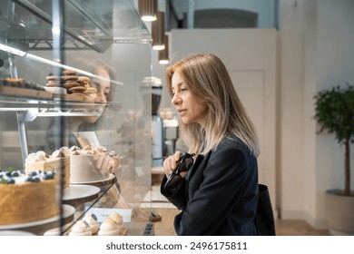 Focused middle aged woman entrepreneur choosing desserts in modern bakery for coffee break near office. Interested businesswoman sweet cake lover looking at cafe case with tasty brownies after lunch. - Powered by Shutterstock