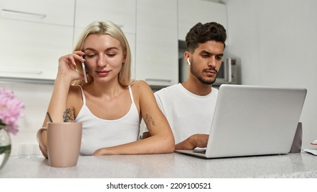 Focused Middle Eastern Man Working On Laptop While European Woman Talking On Smartphone At Table At Home Kitchen. Young Multiracial Couple. Remote Work And Freelance. Domestic Lifestyle. Spacious Flat