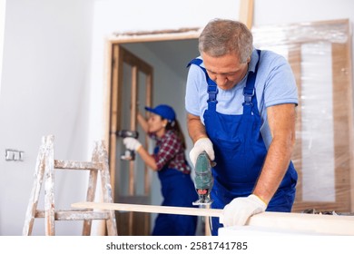 Focused middle-aged man in a blueuniform using a jigsaw machine on wooden plank at home