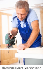 Focused middle-aged man in a blueuniform using a jigsaw machine on wooden plank at home