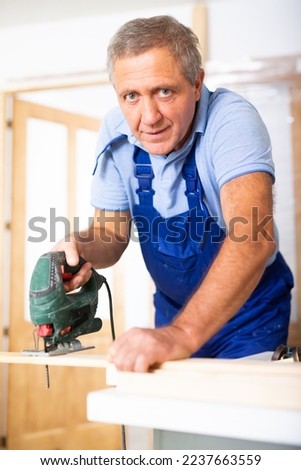 Focused middle-aged man in a blueuniform using a jigsaw machine on wooden plank at home