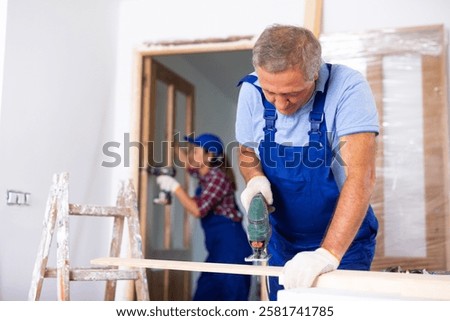 Focused middle-aged man in a blueuniform using a jigsaw machine on wooden plank at home