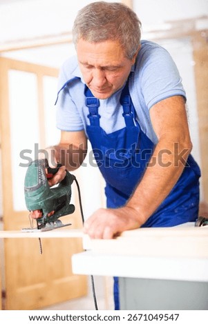 Focused middle-aged man in a blueuniform using a jigsaw machine on wooden plank at home