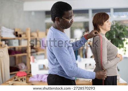 Focused middle-aged tailor carefully taking measurements from female client in sewing studio, preparing to create custom clothing piece with proper fit