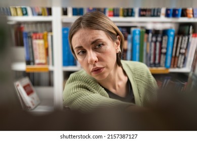 Focused Middle-aged Woman In Bookstore Choosing Book While Walking Alongside Rows Of Bookshelves. Closeup Of Female Student Doing Research In Library, Getting Second Degree. Education Has No Age Limit