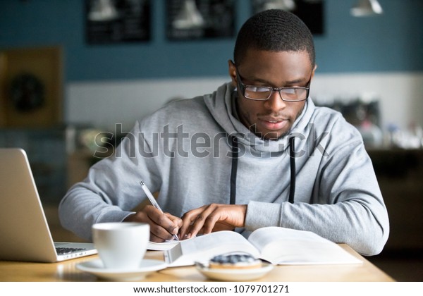 検査や試験の準備をするカフェの本から情報を書き留めたメモを作る眼鏡をかけた 注目を集めたアフリカ系アメリカ人の学生 若い真面目な黒人 コーヒーハウスで勉強または働く若い男性 の写真素材 今すぐ編集