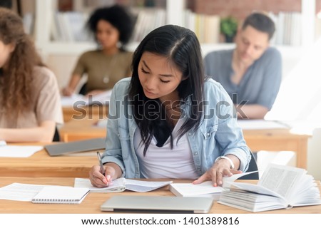 Focused millennial Asian female student sit in shared coworking space studying with handbook writing do homework, concentrated teen girl make notes in workbook prepare for test or exam in library