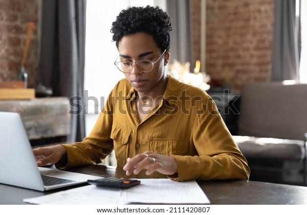 Focused\
millennial Black business woman calculating finance, money, using\
calculator, laptop computer at home workplace table, counting\
budget, paying bills, taxes, rent, mortgage\
fees