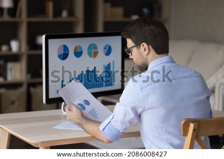 Focused millennial business man analyzing marketing reports on desktop monitor, reviewing paper graphs, financial stats, startup project infographics. Marketer working from home office. Back view
